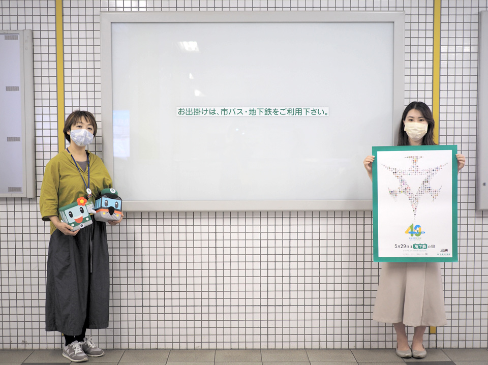 写真：空の電照広告の前に立つ担当者たち。手にマスコットや地下鉄40周年のポスターを掲げている。